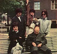 On a bench in a park sit five men, two seated and three standing behind the bench. Clockwise from left is a man in a black suit with Khaki pants; a man in a black suit with black pants; a man wearing a brown coat with Khaki pants and raising his arm jokingly as if to stab the sleeping man below him; a man, wearing a grey/light green coat and Khaki pants and holding out a hat above the sleeping man's head. The sleeping man is wearing green.