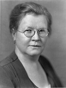 A middle-aged white woman with dark hair, wearing glasses and a dark top