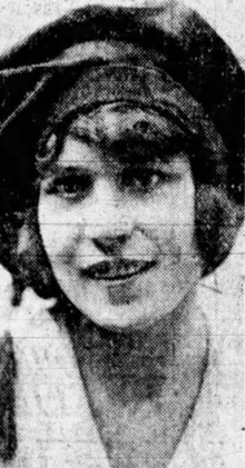 A smiling young white woman with dark hair, wearing a dark hat and a light-colored top