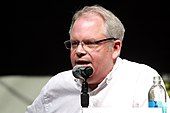 A Caucasian man wearing a white shirt speaks into a microphone.