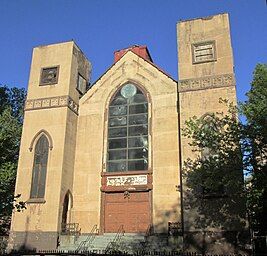 The front of a three-story building is visible. It shows two rectangular towers, one on each side of a recessed bay, all clad in tan stucco. The towers have pointed arched windows on the bottom and square ones on top. The bay has four wooden doors at the bottom and a sign with Hebrew writing on top of them, surmounted by large arched multi-paned window. Atop the roof of the bay is a small metal Star of David.