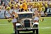 A 1930 Ford Model A Sports coupe painted gold being driven on a football field with three cheerleaders on each side and two additional cheerleaders and a hornet mascot in the back.