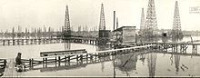 A black-and-white photograph showing a field of oil derricks and thin wooden boardwalks built over the water.