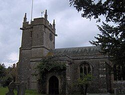 Stone building with square tower at left hand end. In the foreground are trees and gravestones.