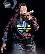 Missy Elliott posing on a grey background wearing a black shirt and gold jewelry.