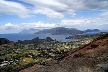 A photograph of the Aeolian Islands, showing Lipari in the middle, Salina to the left, and Panarea to the right. Overhead is a blue but cloudy sky.