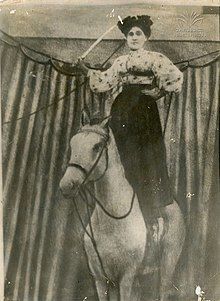 Photograph of a Gurian woman, dressed in a Victorian-style skirt and blouse, standing upon the back of a horse, with a raised sword in her right hand and her left hand on her hip.