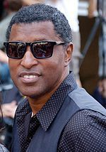 A black man appears in white and black polka dot shirt, black waistcoat and sunglasses