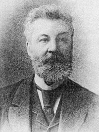 Old grainy photograph of the head and shoulders of a bearded man wearing a three-piece suit, tie and high collar