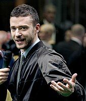 A young white man with stubble, wearing a jacket, blue shirt and tie, being interviewed.