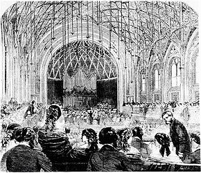 Interior of a concert hall with patrons taking their seats in the foreground, with organ and performers in the background.