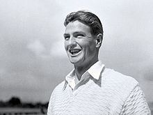 Young man with dark hair parted in the middle, grinning and wearing a white open collar shirt and a white jumper