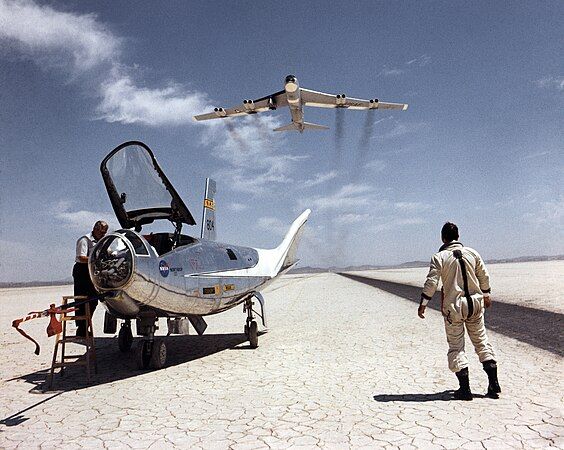 NASA research pilot Bill Dana takes a moment to watch NASA's NB-52B cruise overhead after a research flight in the HL-10. On the left, John Reeves can be seen at the cockpit of the lifting body.