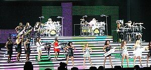 A musical group of women, composed of at least eighteen of them, is performing a song on stage. They play drum kits, keyboards and saxophones, while other females sing and dance.