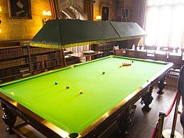 A full-size snooker table in a brightly lit room with bookcases and a boardroom table in the background, all cordoned off at the right-hand side as part of an English country house display