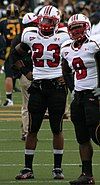 Two football players in complete gear standing side-by-side with their hands on their hips. Both are wearing black pants, white jerseys and white helmets with red face mask.