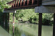 A narrow bridge, supported by sets of cylindrical piers topped with concrete, crosses a muddy river. A much smaller stream, nearly obscured by vegetation, enters the river near one end of the bridge.