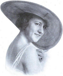 A smiling white woman with bobbed hair, glancing over her shoulder, wearing a large brimmed hat and a dress with bare arms and upper back