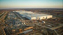 Photo is of a large scale manufacturing facility under construction, captured from above with aerial photography. There are many cranes, heavy equipment, materials, and other machinery surrounding the tall plain walls of the main facility.