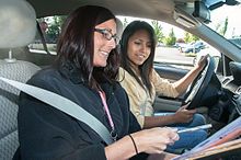 Two women sit in a car. The passenger holds a paper so the driver can see it.