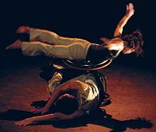 a dancer stretched out balances on another dancer's wheelchair who is curled over, they are on-stage with theatre lights highlighting the dance