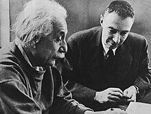 Einstein writing at a desk. Oppenheimer sits beside him, looking on.