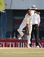 A man in Test cricket whites, on his follow through after releasing the ball while bowling; three stumps and a man in white pants, white shirt and hat are in the background.