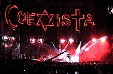 A view of the stage being filmed, with "COEXISTA" spelled out on the video screen, using an Islamic crescent as a "C", a Star of David as an "X", and a Christian cross at a "T". A camera in the middle shows the concert being filmed.