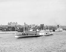 A white steam ship is seen near the shore of the Hudson River in front of the downtown area of Albany; the New York State Capitol can be seen in the background.