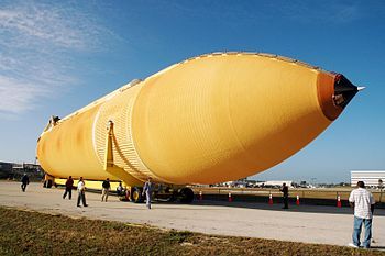 The newly redesigned External Tank turns the corner of the Launch Complex 39 Area Turn Basin parking area on its way to the Vehicle Assembly Building, seen at right.