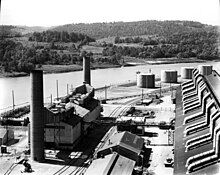 A large dark-coloured rectangular building and a smaller building with three smokestacks. In the background is the river. The steam plant is a small building with two smokestacks.