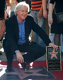 James Cameron, a white man with white hair in a navy suit, kneeling down over his star on the Hollywood Walk of Fame and holding an award