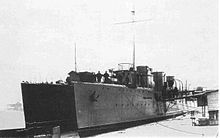 A black-and-white photo shows two ships moored parallel to each other at the pier. On the deck of the nearer ship, two people are standing by the railing. Several people are boarding the ship via a gangway. A section of a low, wooden building is visible on the pier.