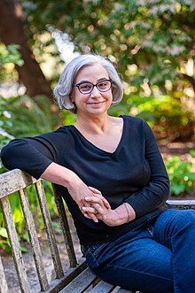 Laurie Frankel sits on a bench in a park. She has short white hair, and wears glasses.