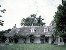 A two-story, white, Dutch Colonial-style house with three dormer windows and shrubbery obscuring the front porch columns.