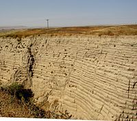 This photo shows another canyon cut into the surrounding flat soil with about 30 distinct horizontal layers of sediment, each clearly demarked from the layer below. Above the canyon a telephone pole can be seen in the distance – the pole provides the perspective that helps the viewer establish that the cut is 30–40 ft (9.1–12.2 m) deep. In the foreground one observes the near edge of the canyon, which help one establish that the canyon is quite narrow and steep walled.