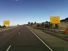 A highway near the top of a ridge. On either side of the highway are big yellow signs reading, "Trucks, Don't be Fooled—4 more miles [6.4 km] of steep grades and sharp curves".