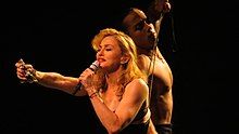 A blonde woman sings to a microphone and has her arm stretched out; behind her, a male pulls a string attached to her garment.