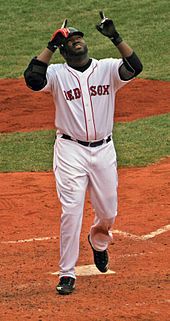 A dark-skinned man in a black baseball jersey and gray pants takes a right-handed baseball swing with a crowd in the background, several people wearing red.