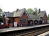 Two sets of rails between platforms beyond which is a brick building with two gables and a canopied area between them; a sign to the left says "Widnes"