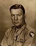 Head and shoulders of a stern-faced white man. A row of ribbon bars are on his left breast as is the Combat Infantryman's Badge and the star-shaped Medal of Honor hangs from a ribbon around his neck.