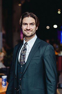 Colour photograph of Hendrik Johannes Terras aged 29. He is wearing an emerald greed suit with a white shirt and a patterned tie. he appears to be at an event, though the background is blury. he has a short, stubbly beard and a welcoming smile.