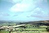 The houses and church of a small village can be seen in the bottom left of the picture. It is surrounded by a patchwork of fields with some trees on a hillside. Large hills in the distance.