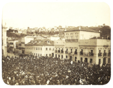 An old photograph showing a crowded square in front of a large, white, multi-storied building