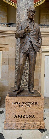 A brown bronze statue of Barry Goldwater. He is leaning left, and his right hand is casually reached out while his right elbow is still at his side. He is wearing a suit, and on the pedestal below the statue is the text "BARRY GOLDWATER (1909-1998), ARIZONA".