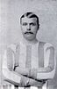 Monochrome photo of head and upper torso of young man with shiny side-parted hair and large moustache, wearing long-sleeved sports shirt with vertical stripes