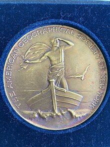 Image of bronze circular medal in dark blue velvet. The medal includes the text "The American Geographical Society of New York" in all capital letters around the edge of the medal. The center image on the medal is a man standing on a boat looking to his left with a bird on the left.