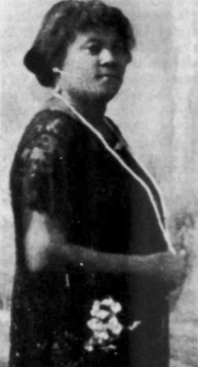 A Black woman with dark hair cut in a bob with bangs, wearing a dark dress and a long strand of beads or pearls