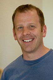 A red-haired man wearing a blue T-shirt looking directly forward and smiling.