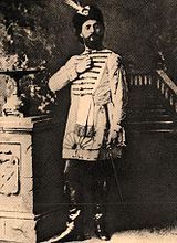 Monochrome photograph of a man standing, wearing leather books, knee-length tunic and feathered hat